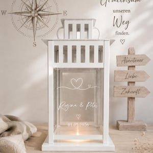 Pode incluir: Um lampião de metal branco com painéis de vidro apresentando um design gravado em branco de um coração, os nomes Regina & Peter e a data 01.05.2026. O lampião possui uma alça superior quadrada e uma pequena luz brilhando na base.