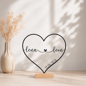 Könnte beinhalten: Schwarze herzförmige Dekoration aus Metall mit den Namen "lena" und "leon" sowie dem Datum "11.06.2026". Das Herz steht auf einem kleinen Holzsockel. Im Hintergrund ist eine Vase mit Trockenblumen zu sehen.