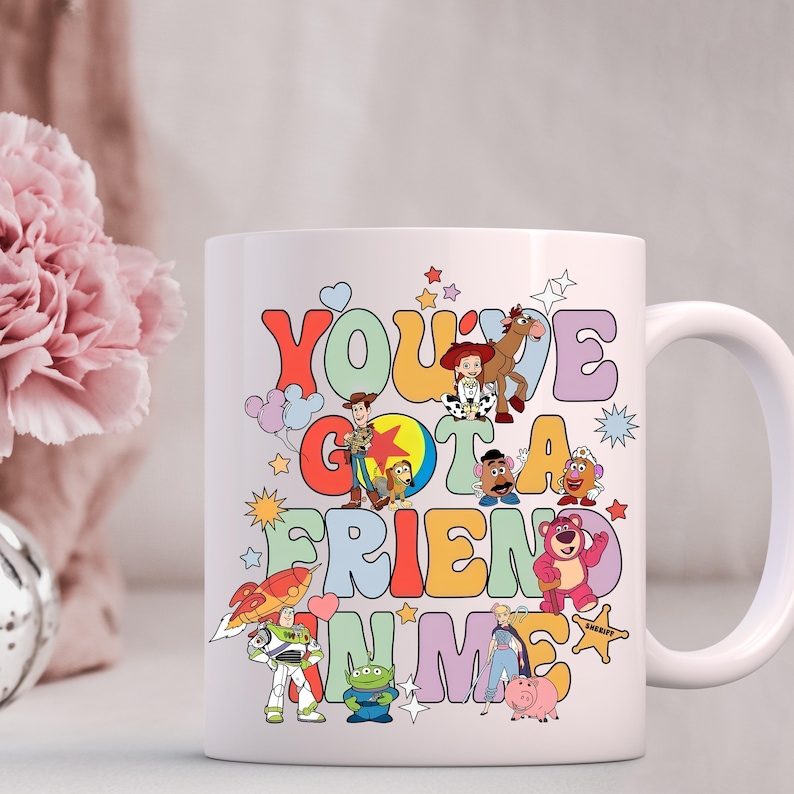 K&ouml;nnte beinhalten: Wei&szlig;e Keramik-Kaffeetasse mit einer bunten Grafik von Figuren aus dem Toy Story-Film-Franchise. Der Text auf der Tasse lautet "You've Got a Friend in Me".