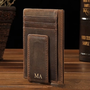 May include: A dark brown leather card holder featuring a vertical money clip on the front. The exterior surface displays visible stitching along the edges and includes the embossed gold-colored letters MA at the bottom of the money clip section.