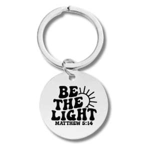 Puede incluir: Un llavero plateado con una etiqueta redonda que dice "Sé la luz" en una fuente retro con un gráfico de sol y "Mateo 5:14" debajo.