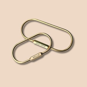 May include: Two gold-colored keychains interlocked. One keychain has the word "BELLA" engraved on it, and the other has "your name" engraved. The keychains are oval-shaped with a connecting bar.
