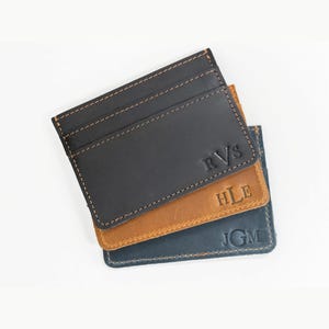 May include: Three rectangular leather card holders stacked on top of each other. The top holder is black with the embossed letters RVS, the middle is tan with HLE, and the bottom is dark blue with JGM. Each features visible contrast stitching.