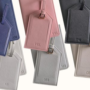 May include: A collection of personalized luggage tags and passport holders in various colors, including navy blue, pink, gray, and cream. Each tag features initials and a matching passport holder. The tags have a buckle closure.