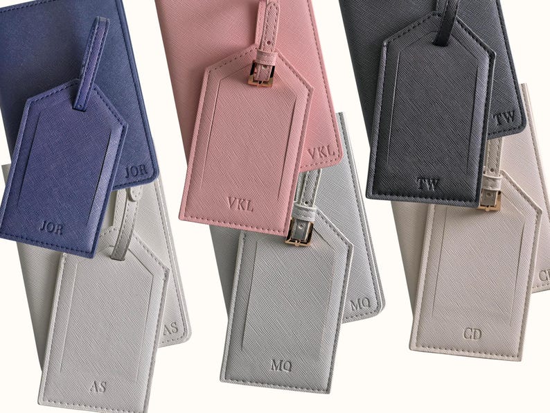May include: A collection of personalized luggage tags and passport holders in various colors, including navy blue, pink, gray, and cream. Each tag features initials and a matching passport holder. The tags have a buckle closure.
