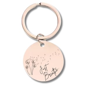 Puede incluir: Un llavero de oro rosa con un colgante redondo. El colgante presenta un diseño de diente de león con las palabras "Just Breathe" en una fuente manuscrita. El llavero tiene un anillo plateado.