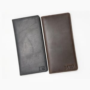 Könnte beinhalten: Zwei rechteckige Scheckbuchhüllen aus Leder, eine in Schwarz und eine in Dunkelbraun. Jede Hülle hat eine glatte, matte Oberfläche mit sichtbarer natürlicher Maserung und ist in der unteren Ecke mit zweibuchstabigen Initialen geprägt, KS und MW.