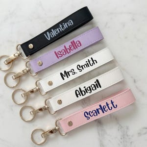 Könnte beinhalten: Fünf Schlüsselanhänger in Schwarz, Lavendel, Weiß und Rosa, jeweils mit einem goldfarbenen Metallverschluss und Ring. Die Bänder sind mit den Namen Valentina, Isabella, Mrs. Smith, Abigail und Scarlett in verschiedenen Schriftfarben personalisiert.