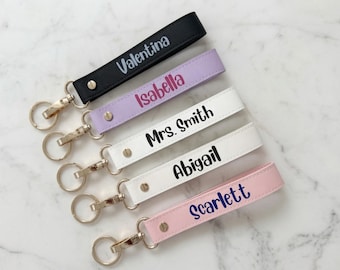 May include: Five wristlet keychains in black, lavender, white, and pink, each featuring a gold-tone metal clasp and ring. The straps are personalized with the names Valentina, Isabella, Mrs. Smith, Abigail, and Scarlett in various colored fonts.
