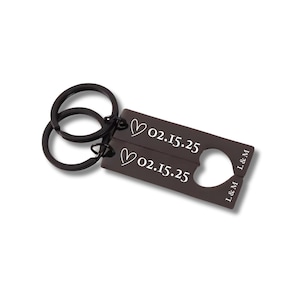 Puede incluir: Un llavero de metal marrón oscuro con dos llaveros y una etiqueta rectangular. La etiqueta tiene dos corazones, la fecha "02.15.25" y las iniciales "L&M" grabadas. El llavero está sobre un fondo blanco.
