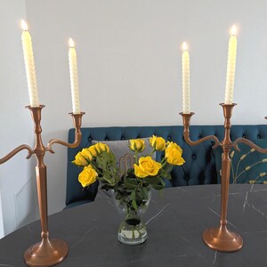 Peut inclure: Deux candélabres cuivrés, chacun avec trois bougies torsadées blanches allumées. Un vase de roses jaunes est placé entre eux. Les candélabres sont sur une table sombre, avec un fond bleu rembourré.