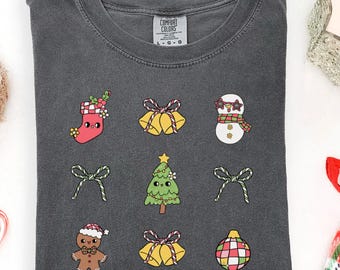 May include: A charcoal grey t-shirt featuring embroidered festive designs: a stocking, bells, a snowman, bows, a Christmas tree, a gingerbread man, and an ornament. A reindeer plush, a candy cane, a candle, and plaid pyjama bottoms are also visible.
