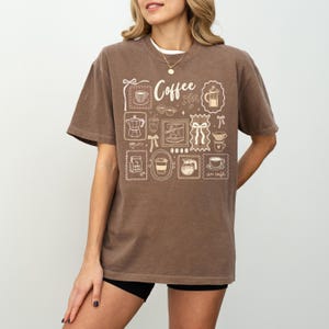 May include: A brown t-shirt with a white graphic design featuring coffee-related illustrations and the word "Coffee". The design includes images of coffee cups, coffee makers, and other coffee-themed elements.
