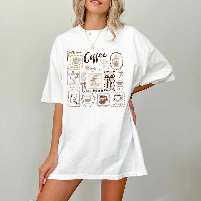 May include: White t-shirt with a brown coffee-themed graphic. The design includes various coffee-related illustrations such as a coffee cup, coffee maker, and the word "Coffee". The shirt has a relaxed fit.