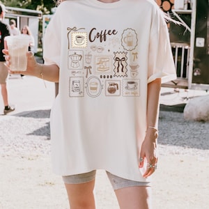 May include: Cream-colored t-shirt featuring a coffee-themed design. The design includes various coffee-related illustrations such as coffee cups, a coffee maker, and the word "Coffee". The shirt has short sleeves and a relaxed fit.