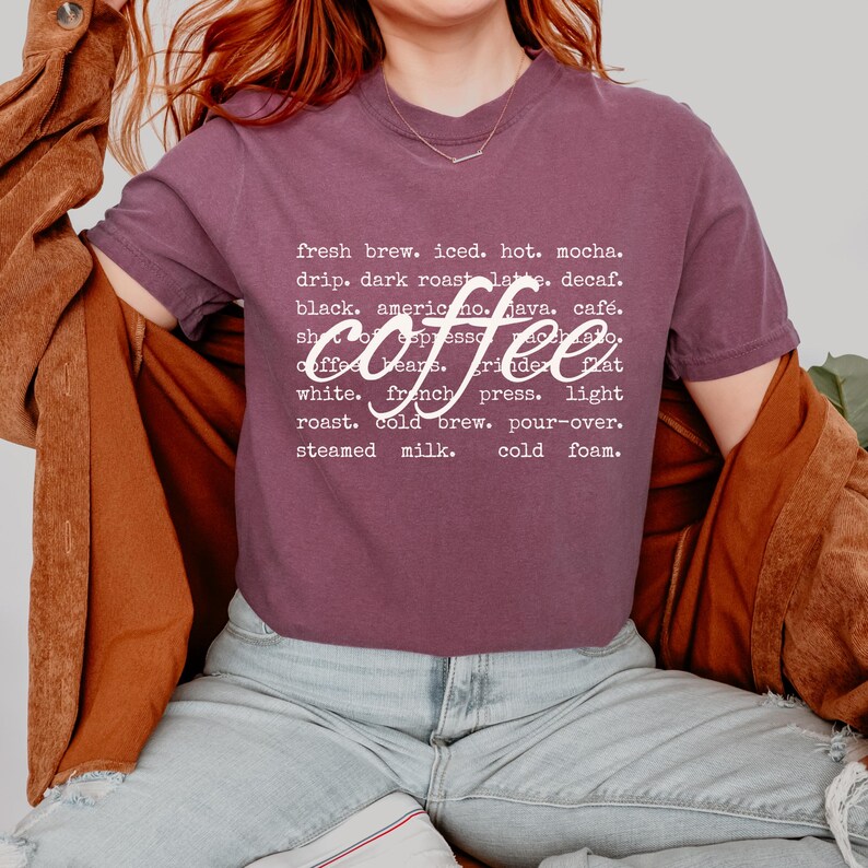 May include: A mauve t-shirt with the word "coffee" in a stylized font. The shirt features a list of coffee-related terms in white text, including "fresh brew," "iced," and "mocha."