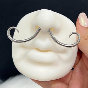 May include: A novelty nose and mouth prop made of white material, featuring a pair of silver-colored mustache-shaped nose rings. The prop is designed to resemble a face with a mustache.