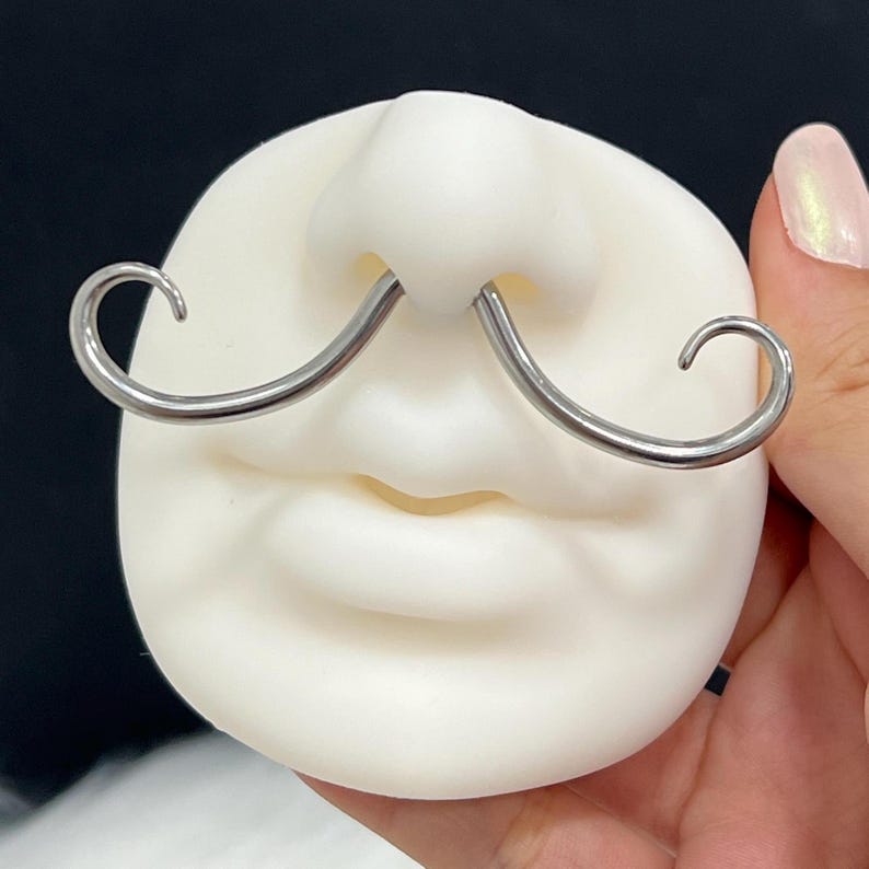 May include: A novelty nose and mouth prop made of white material, featuring a pair of silver-colored mustache-shaped nose rings. The prop is designed to resemble a face with a mustache.