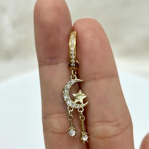 Könnte beinhalten: Goldener Creolenohrring mit einem baumelnden Halbmond-Anhänger mit einem Stern und zwei baumelnden Sternen. Der Anhänger ist mit klaren Zirkonia-Steinen besetzt.