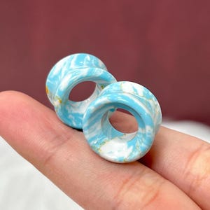 May include: Two blue and white marbled ear gauges. The gauges have a tunnel design and are held in place by a finger. The gauges have a swirl pattern of light blue and white. The gauges are made of a glossy material.