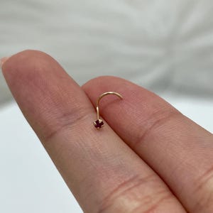 Puede incluir: Un piercing de nariz de color dorado con una pequeña gema redonda de color rojo oscuro. El piercing de nariz tiene forma curva con una pequeña piedra decorativa en el extremo. La joya se muestra sobre un fondo borroso.