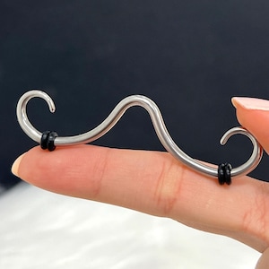 May include: A silver-colored, mustache-shaped body jewelry piece. The curved design features a central wave with rounded ends, each with a small black rubber ring. The jewelry is held by a finger against a dark background.
