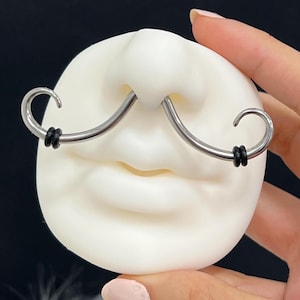 May include: A silver-colored, curved nose ring with black rubber rings on each end, shaped like a mustache, is displayed on a white, face-shaped object. The nose ring is centered on the nose of the face.