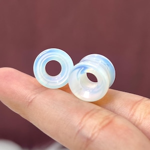 May include: Two opalescent ear gauges with a swirling blue and white pattern. These body jewelry pieces have a cylindrical shape with a hollow center. The gauges are designed to stretch ear piercings. The background is a solid maroon color.