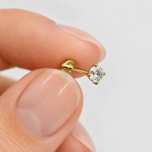 May include: A gold-tone flat back stud earring featuring a single round clear gemstone held in a four-prong setting. The post is straight and smooth, extending from the back of the setting to a small, circular flat disc base at the end.