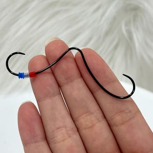 Puede incluir: Un bigote de alambre negro con una cuenta roja, blanca y azul en el centro.