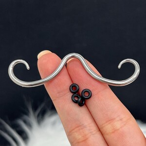 May include: A silver-colored mustache-shaped piercing jewelry with curved ends. Several small black rubber rings are also present. The jewelry is held in a hand against a dark background.