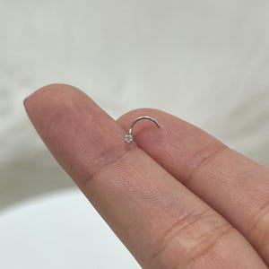 May include: A small, curved nose ring with a clear, round gemstone at the end. The jewelry is silver-colored and is shown on a finger against a neutral background. The gemstone appears to be approximately 0.08 inches in diameter.