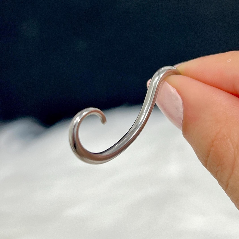 May include: A silver-colored, curved metal object, held by a person. The object has a smooth, polished surface and a flowing, abstract shape. The design is reminiscent of a stylized letter or a decorative element.