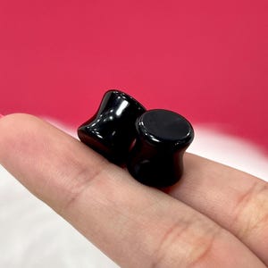 Puede incluir: Dos dilatadores de oreja negros con un diseño acampanado. Los dilatadores están hechos de un material liso y brillante y se muestran sobre un fondo rosa. Los dilatadores de oreja son un tipo de joyería de modificación corporal.