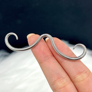 May include: A silver-colored mustache-shaped nose ring. The nose ring is made of a curved, smooth metal with rounded ends. The design is a playful take on facial hair, suitable for a unique style statement.