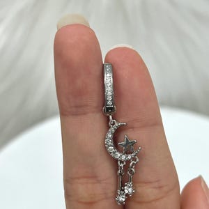 May include: Silver hoop earrings with a crescent moon and star charm dangling from the bottom. The charm is adorned with small clear crystals.