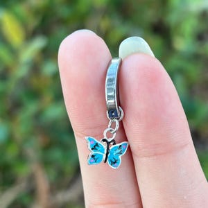Pode incluir: Um brinco de argola prateado com um pequeno pingente de borboleta pendurado. A borboleta tem asas azuis brilhantes com detalhes em rosa e preto. O brinco é segurado entre dois dedos contra um fundo verde desfocado.