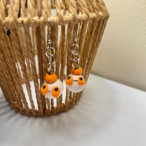 Può includere: Un paio di orecchini pendenti con ciondoli a forma di zucca in argilla bianca. Ogni zucca ha occhi arancioni e uno stelo.