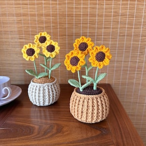 Op de afbeelding: Twee gehaakte zonnebloemarrangementen in decoratieve potten. De zonnebloemen hebben gele bloemblaadjes en bruine centra, met groene stelen en bladeren. Eén pot is beige, de andere is lichtbruin, beide met een geribbelde textuur. De opstelling staat op een houten oppervlak.