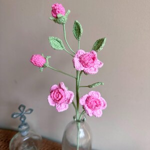 K&ouml;nnte beinhalten: Ein Strau&szlig; geh&auml;kelter rosa Rosen mit gr&uuml;nen Bl&auml;ttern in einer durchsichtigen Glasvase.