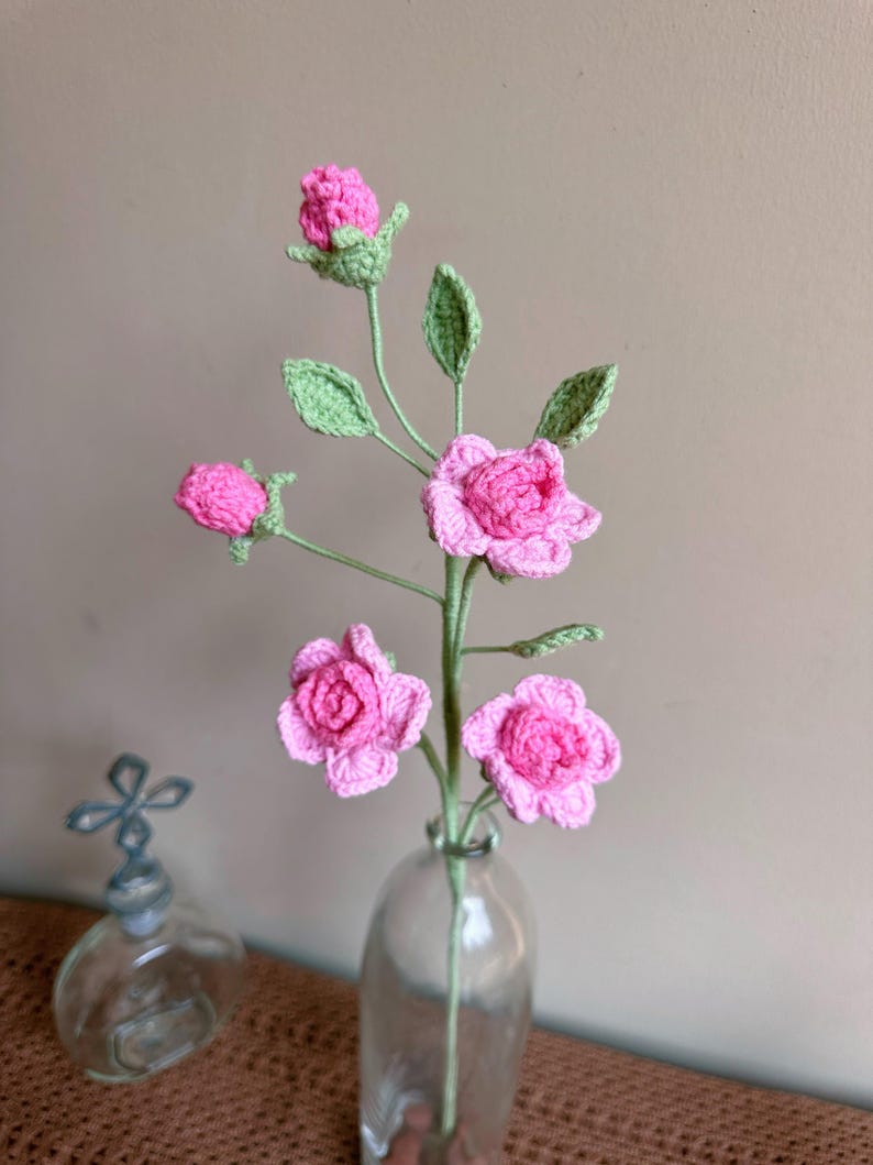 K&ouml;nnte beinhalten: Ein Strau&szlig; geh&auml;kelter rosa Rosen mit gr&uuml;nen Bl&auml;ttern in einer durchsichtigen Glasvase.