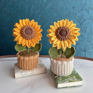 May include: Two crochet sunflowers in pots. The sunflowers have yellow petals and brown centers. The pots are beige and white, and the green leaves are visible. The sunflowers are sitting on top of books.