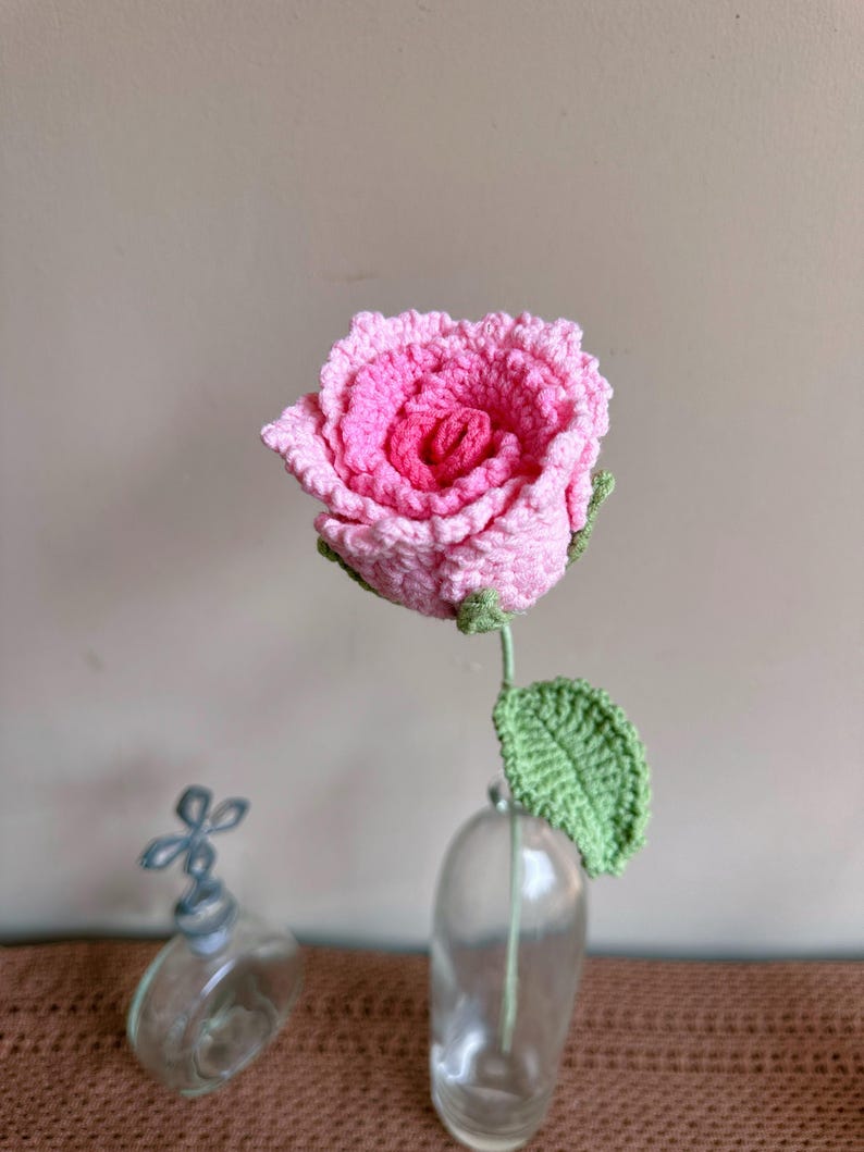 K&ouml;nnte beinhalten: Eine geh&auml;kelte rosa Rose mit gr&uuml;nen Bl&auml;ttern in einer durchsichtigen Glasvase. Die Vase steht auf einer braunen Oberfl&auml;che.