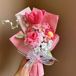 K&ouml;nnte beinhalten: Ein geh&auml;kelter Strau&szlig; aus rosa und wei&szlig;en Blumen, darunter eine Rose, eine Tulpe und G&auml;nsebl&uuml;mchen, in rosa Seidenpapier verpackt und mit einem wei&szlig;en Band gebunden.