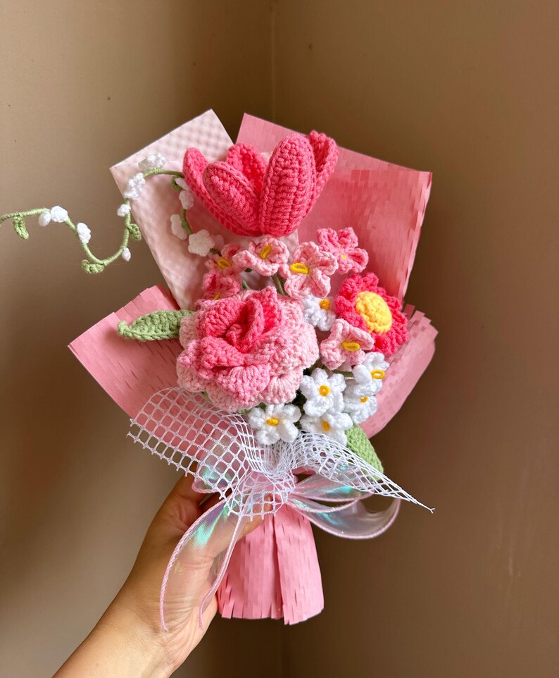 K&ouml;nnte beinhalten: Ein geh&auml;kelter Strau&szlig; aus rosa und wei&szlig;en Blumen, darunter eine Rose, eine Tulpe und G&auml;nsebl&uuml;mchen, in rosa Seidenpapier verpackt und mit einem wei&szlig;en Band gebunden.