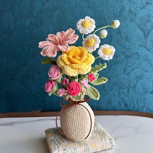 Puede incluir: Un arreglo floral de ganchillo con una rosa amarilla grande, una margarita rosa, margaritas blancas con centro amarillo y pequeñas flores rosas. Las flores tienen hojas verdes y se exhiben en un jarrón beige de ganchillo con un cordón marrón.