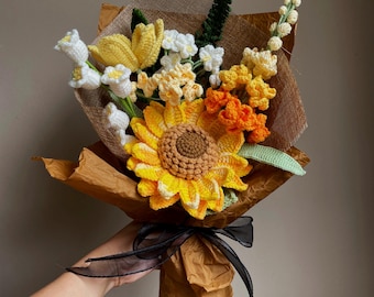 May include: A crochet flower bouquet featuring a large yellow sunflower with a brown center, yellow tulips, orange blossoms, and white bell-shaped flowers. The arrangement is wrapped in brown paper and tied with a black sheer ribbon.