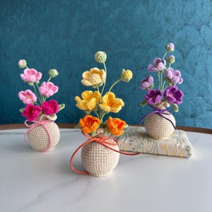 May include: Three crochet flower arrangements in small, beige vases. The flowers are pink, yellow, and purple, with green stems and buds. Each vase is tied with a ribbon. The arrangements sit on a white surface against a teal background.