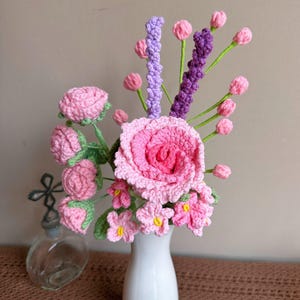 K&ouml;nnte beinhalten: Ein geh&auml;kelter Strau&szlig; aus rosa und lila Blumen in einer wei&szlig;en Vase. Die Blumen sind in einer Vase auf einer braunen Oberfl&auml;che angeordnet.