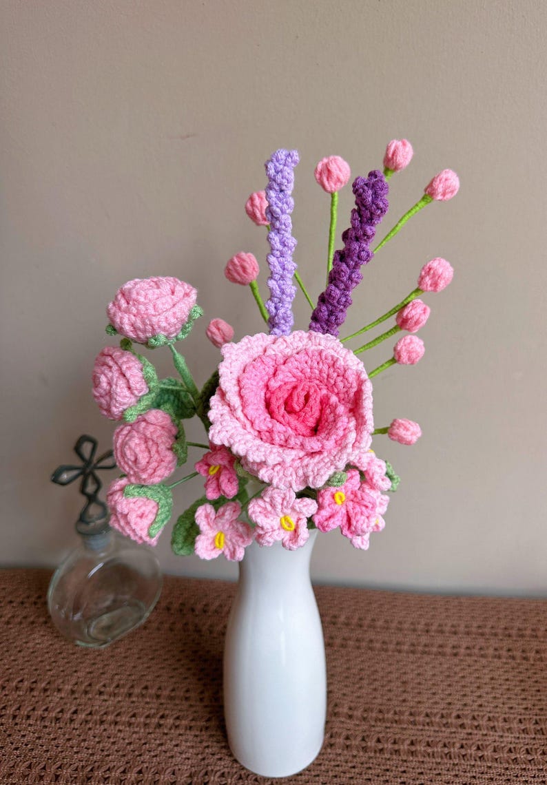 K&ouml;nnte beinhalten: Ein geh&auml;kelter Strau&szlig; aus rosa und lila Blumen in einer wei&szlig;en Vase. Die Blumen sind in einer Vase auf einer braunen Oberfl&auml;che angeordnet.